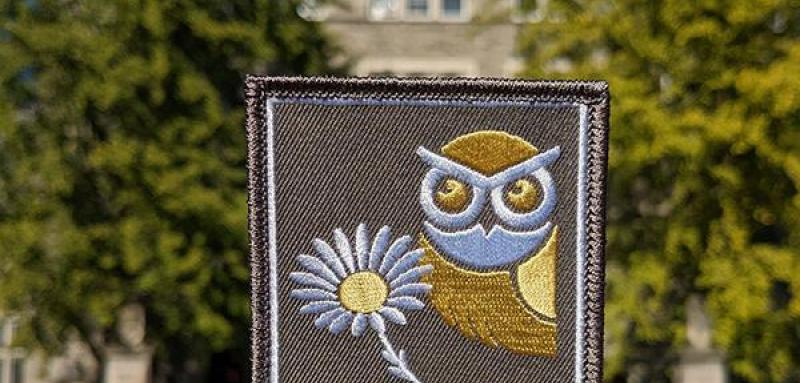 A hand holding an iron-on patch in front of Pem Arch. The patch is gray and features an owl, a daisy, and the words "#BMCGOLD."